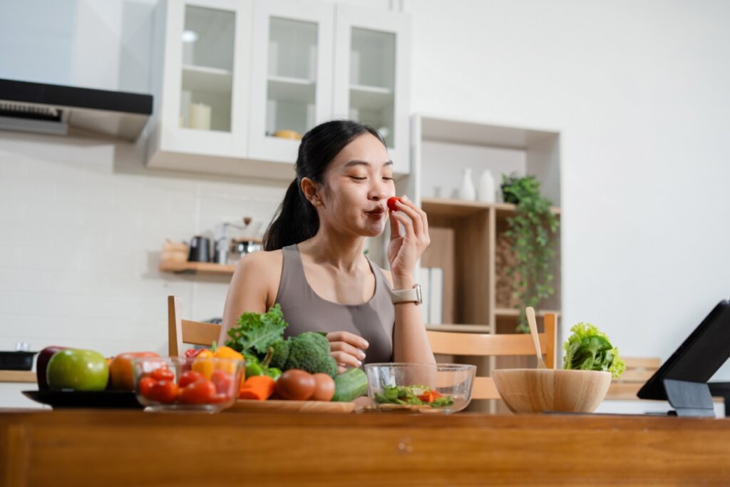 Mulher perto da mesa na cozinha experimentando um tomate enquanto prepara uma refeição com legumes, verduras e frutas dispostos sobre a bancada