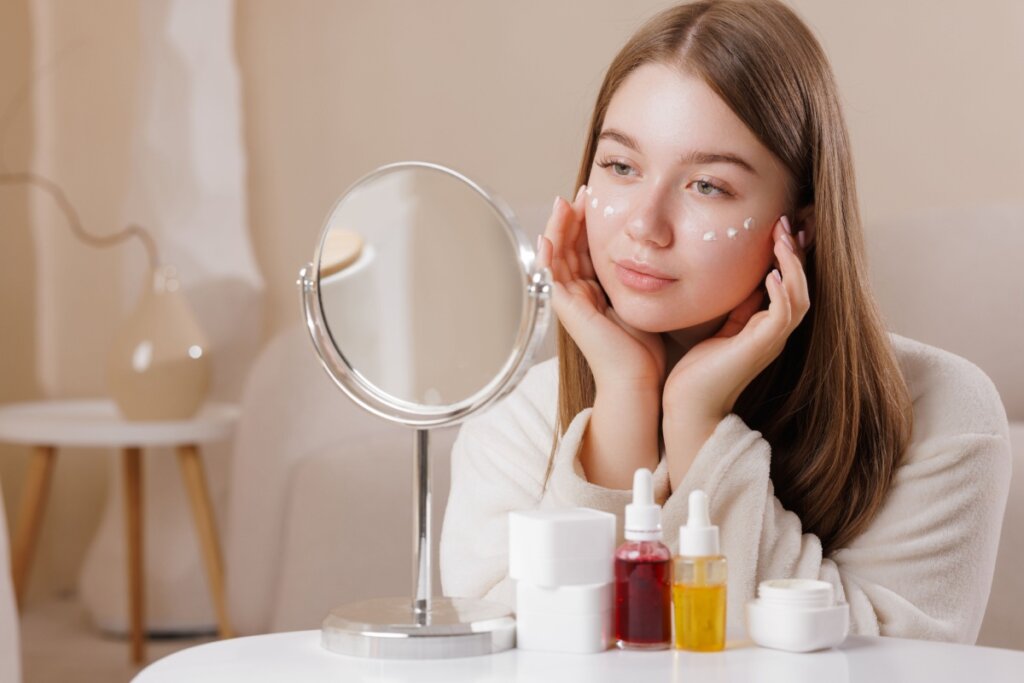 Jovem mulher aplicando pontos de creme de cuidados da pele nas bochechas enquanto olha no espelho redondo. produtos de cuidado com a pele em cima da mesa