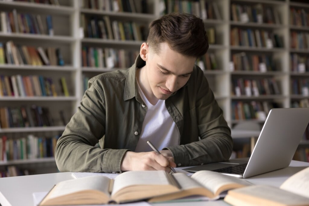 Jovem escrevendo em caderno fazendo resumo, ele tem o cabelo curto e está usando camisa de botões verde-escura e camiseta branca