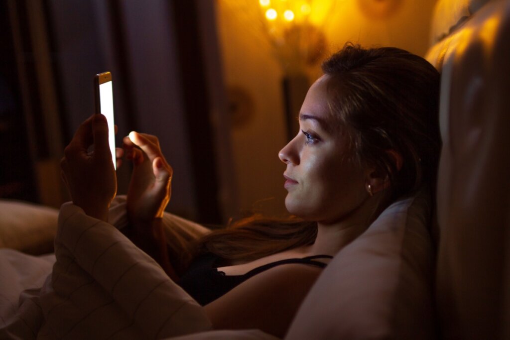 Uma mulher jovem deitada na cama em um quarto escuro, usando um smartphone. A luz brilhante da tela do celular ilumina intensamente o seu rosto e as suas mãos, criando um forte contraste com a penumbra do ambiente.