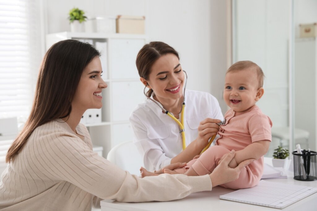 A imagem mostra uma médica examinando um bebê com um estetoscópio, enquanto a mãe observa sorrindo ao lado. O bebê está sentado sobre a mesa de atendimento e aparenta estar tranquilo e feliz. O ambiente é claro e organizado, típico de um consultório pediátrico