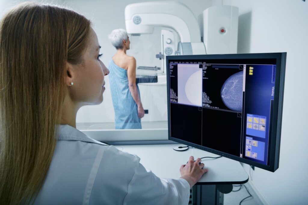 Médica operando máquina de mamografia com paciente realizando o exame ao fundo
