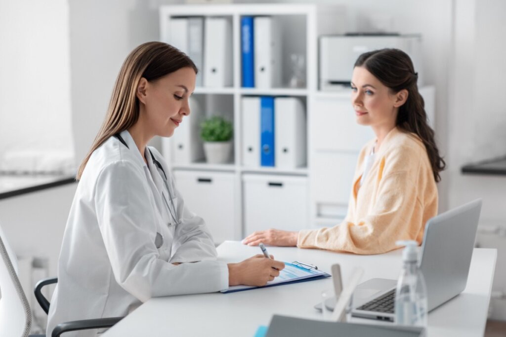 Médica com cabelo liso, solto usando jaleco branco fazendo anotações em prancheta e paciente sentada de frente pra médica com o cabelo liso semi preso e usando camisa de manga longa laranja-clara