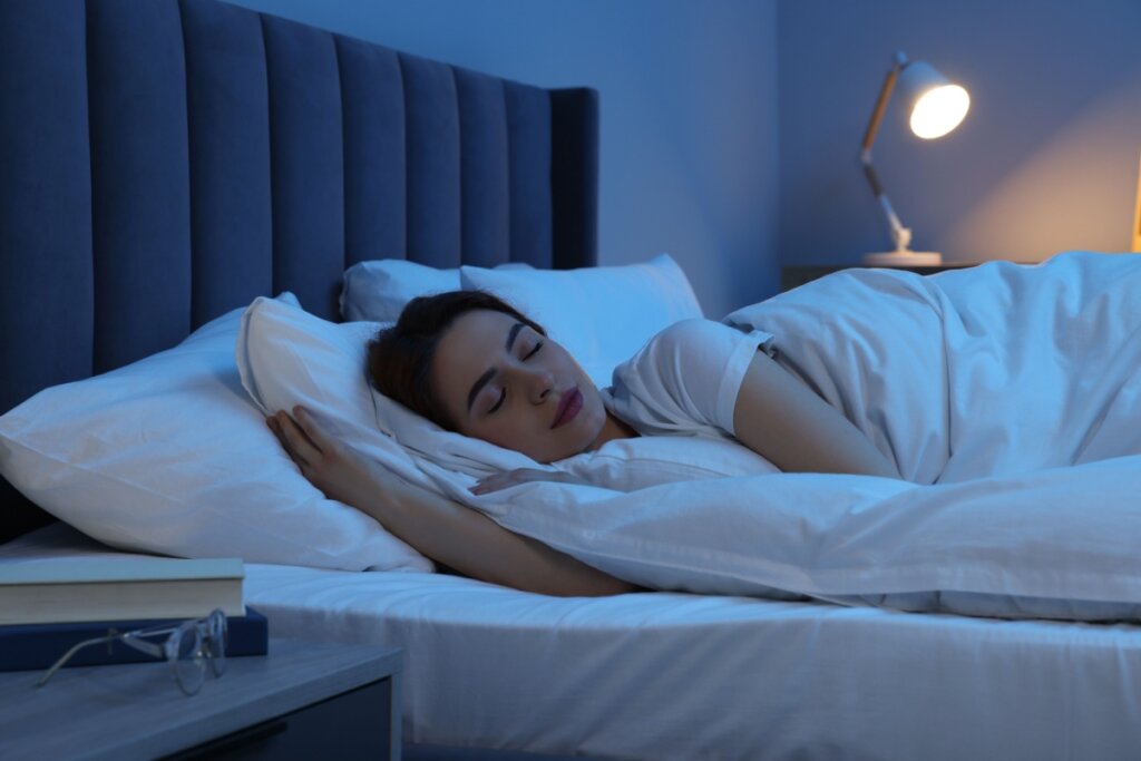 Mulher dormindo em cama, coberta por cobertor branco e usando camiseta branca em quarto escuro