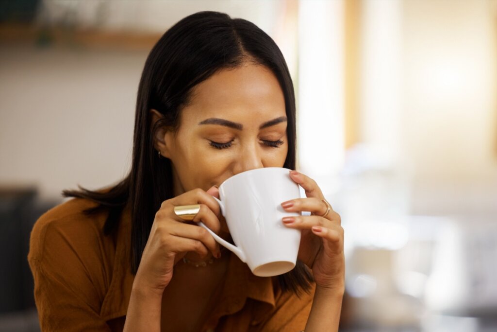 Mulher com cabelo liso preto curto, usando camiseta marrom e tomando chá em caneca branca usando anel dourado em um dos dedos