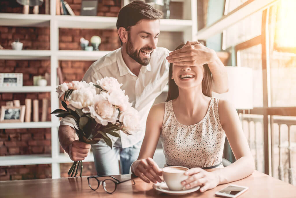 Fotografia em ambiente interno mostrando um homem surpreendendo uma mulher com um presente romântico. Em primeiro plano, a mulher está sentada à mesa, sorrindo alegremente com os olhos cobertos pela mão do parceiro, que está de pé logo atrás dela. Ela segura uma xícara e usa um vestido claro estampado. O homem, sorridente e com barba, veste uma camisa branca de mangas curtas e estende um buquê de flores claras na direção dela com a outra mão. Sobre a mesa de madeira, há também um par de óculos de grau e um smartphone. Ao fundo, vê-se uma estante de livros e uma parede de tijolos aparentes sob uma iluminação quente e difusa.