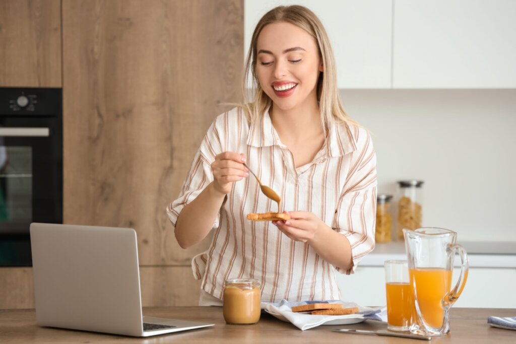 Mulher sorrindo enquanto passa pasta de amendoim em uma torrada na cozinha, ao lado de um notebook e suco