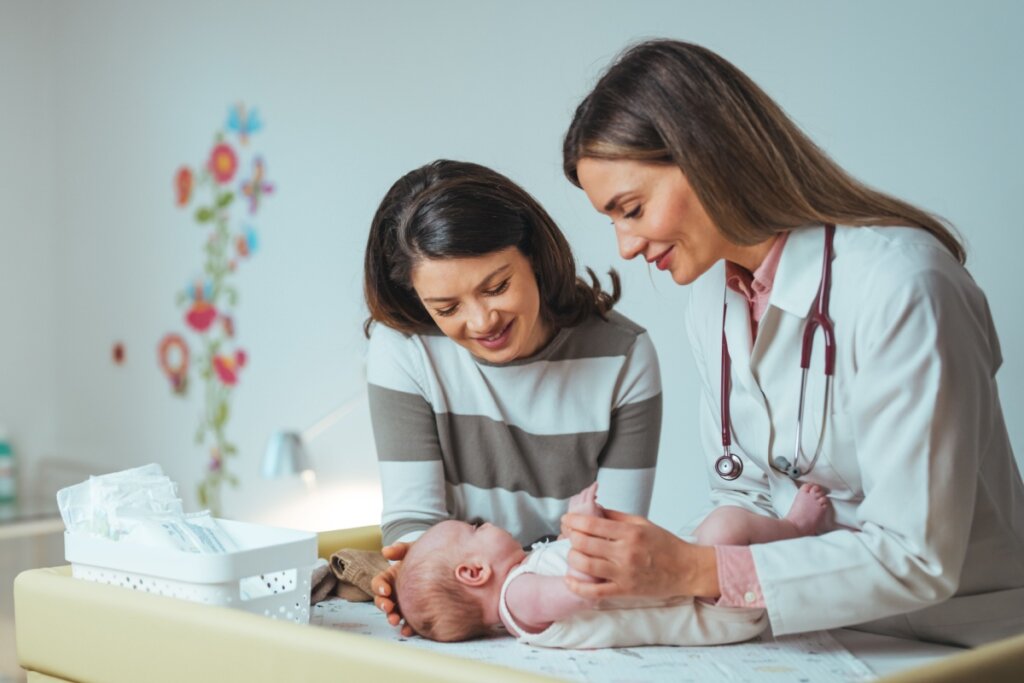 Pediatra com cabelo solto, usando jaleco branco examinando bebê deitado usando macacão branco ao lado da mãe que está com o cabelo solto e usando blusa de manga longa com listras brancas e cinza