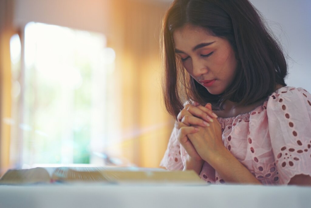 Mulher rezando de olhos fechados e com as mãos unidas no quarto