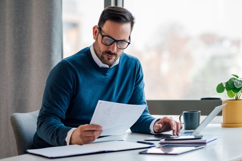 Homem sentado em frente a um notebook e olhando para um papel