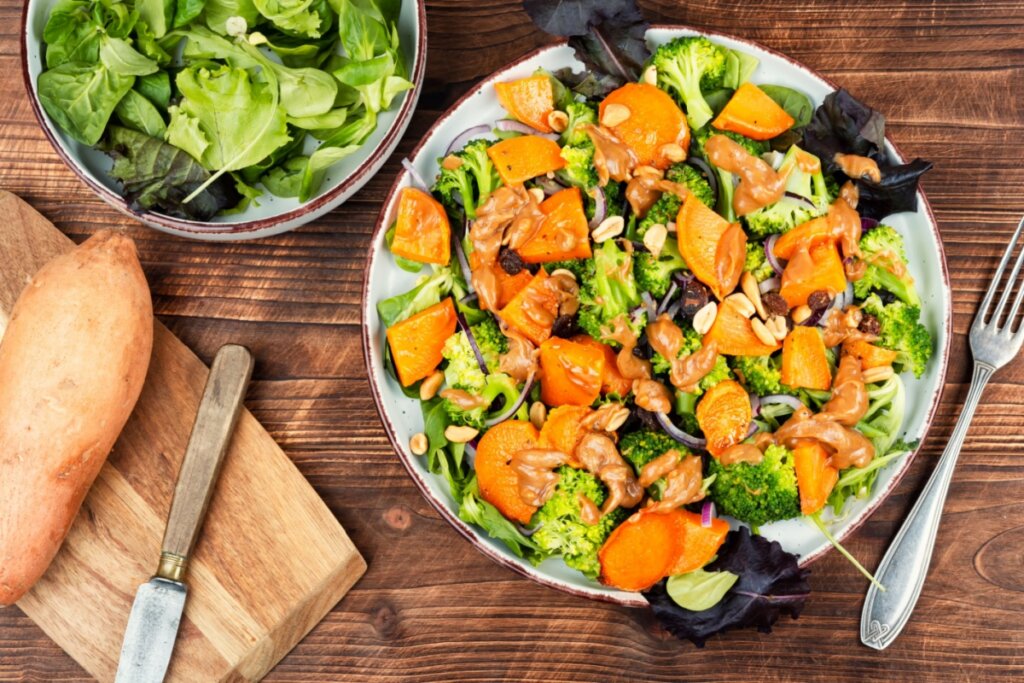 A imagem mostra uma salada colorida preparada com folhas verdes, brócolis, batata-doce em rodelas, cebola roxa, amendoim e molho cremoso espalhado por cima. Servido em um prato sobre uma mesa de madeira