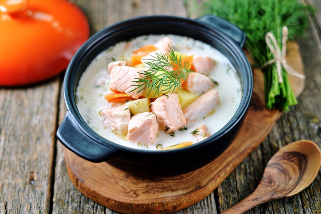 Panela preta com sopa de salmão com batata e cenoura em cima de uma tábua de madeira 