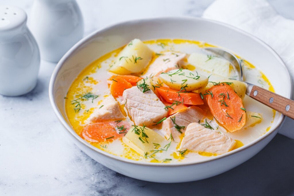 A imagem mostra uma sopa cremosa servida em um prato fundo branco. A receita contém pedaços de salmão, batatas em cubos e rodelas de cenoura mergulhados em um caldo claro e levemente cremoso. A finalização é feita com ervas frescas picadas, que trazem um toque verde ao prato.