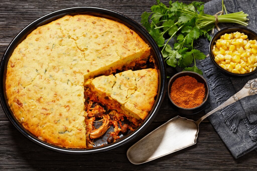 A imagem mostra uma torta de frango com massa dourada à base de milho, servida em uma forma redonda com uma fatia retirada, revelando o recheio de frango desfiado e temperado. Ao lado, aparecem ingredientes como milho, salsa e páprica