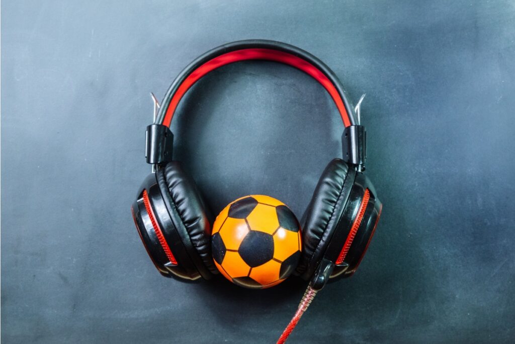 Vista superior de um headphone de haste preta e detalhes vermelhos posicionado sobre uma superfície escura e fosca. No centro do arco dos fones, encaixada entre as almofadas auriculares, está uma pequena bola de futebol nas cores laranja e preta.