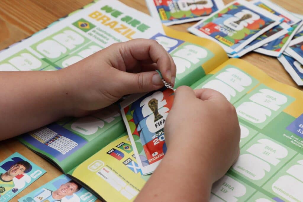Duas mãos aparecem abrindo um pacote de figurinhas oficial da Copa do Mundo de 2026. O pacote está posicionado sobre as páginas abertas de um álbum de figurinhas temático da seleção do Brasil ("We Are Brazil"), com alguns cromos soltos visíveis nas proximidades.