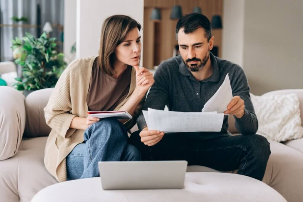 Homem e mulher sentados em um sofá revisando juntos papéis e documentos com atenção, com um notebook aberto sobre uma mesa de centro à frente.
