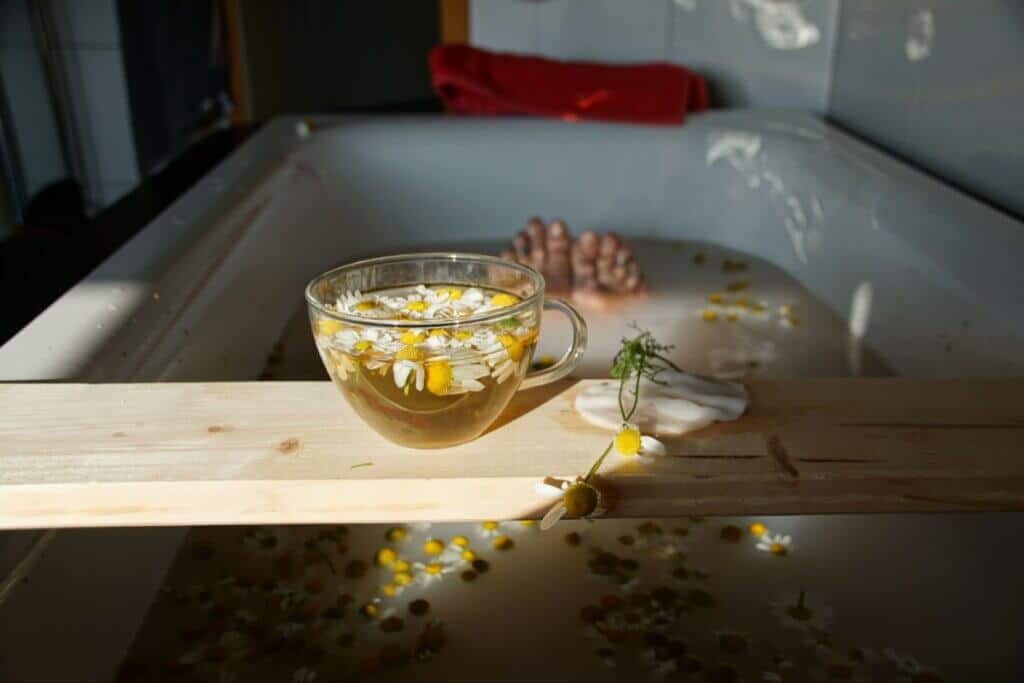 Uma banheira branca cheia de água de aspecto leitoso, com diversas flores de camomila flutuando na superfície. Ao fundo, os pés de uma pessoa de pele clara aparecem submersos na água. Em primeiro plano, uma tábua de madeira clara atravessa a largura da banheira, servindo de suporte para uma xícara de vidro transparente cheia de chá com flores de camomila frescas dentro. Ao lado da xícara, há um sabonete branco e um pequeno ramo da erva