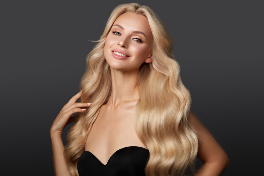 Mulher loira sorrindo com cabelos longos e ondulados, usando um top tomara que caia preto contra um fundo cinza escuro.