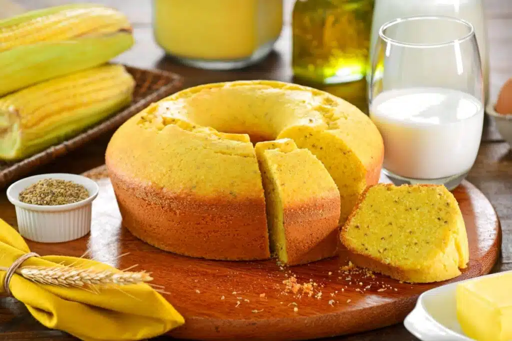 Um bolo de milho dourado e fofinho salpicado com pequenas sementes de erva-doce na massa. O bolo está sobre uma tábua redonda de madeira, com uma fatia cortada e deitada ao lado. Ao redor da tábua, encontram-se espigas de milho verde, um pequeno pote branco com sementes de erva-doce, um guardanapo amarelo, um copo de leite e um tablete de manteiga.