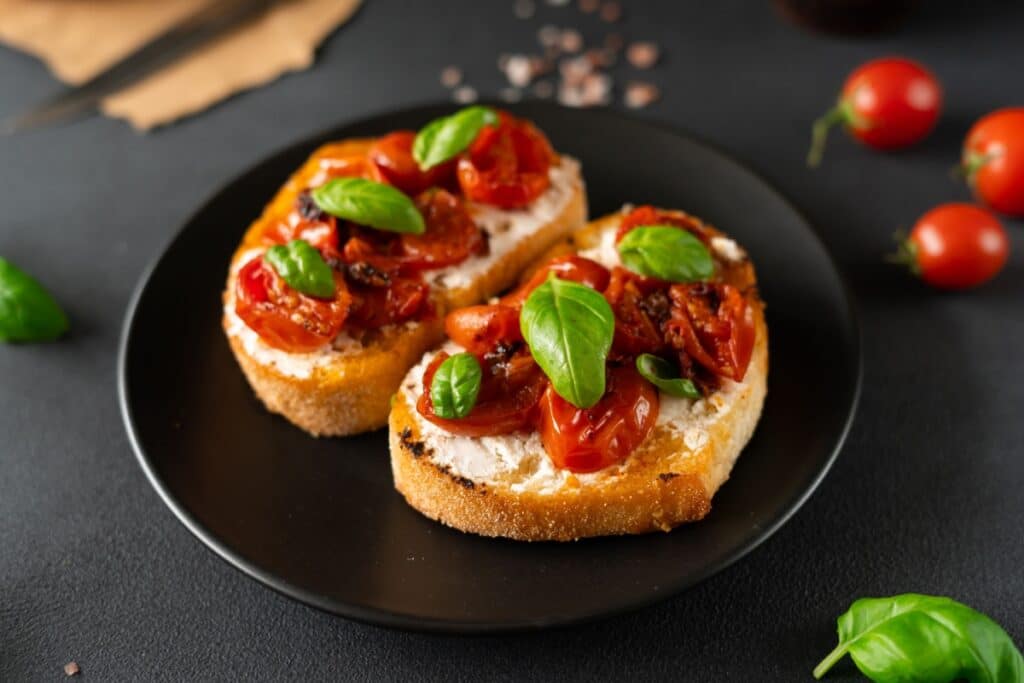 A imagem mostra três bruschettas servidas em um prato preto, preparadas com fatias de pão tostado, uma camada cremosa de queijo branco, tomates-cereja assados e folhas frescas de manjericão.