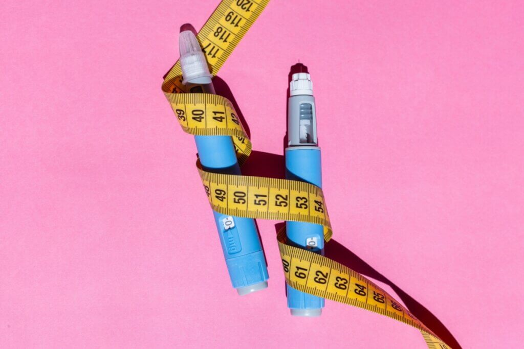 Duas canetas de aplicação de medicamento na cor azul estão posicionadas verticalmente sobre um fundo rosa vibrante. Uma fita métrica amarela envolve as duas canetas de forma sinuosa, criando uma conexão visual entre elas. A iluminação é direta, projetando sombras nítidas à direita dos objetos. A imagem sugere uma relação entre o uso de medicamentos injetáveis e o controle de peso ou medidas corporais.