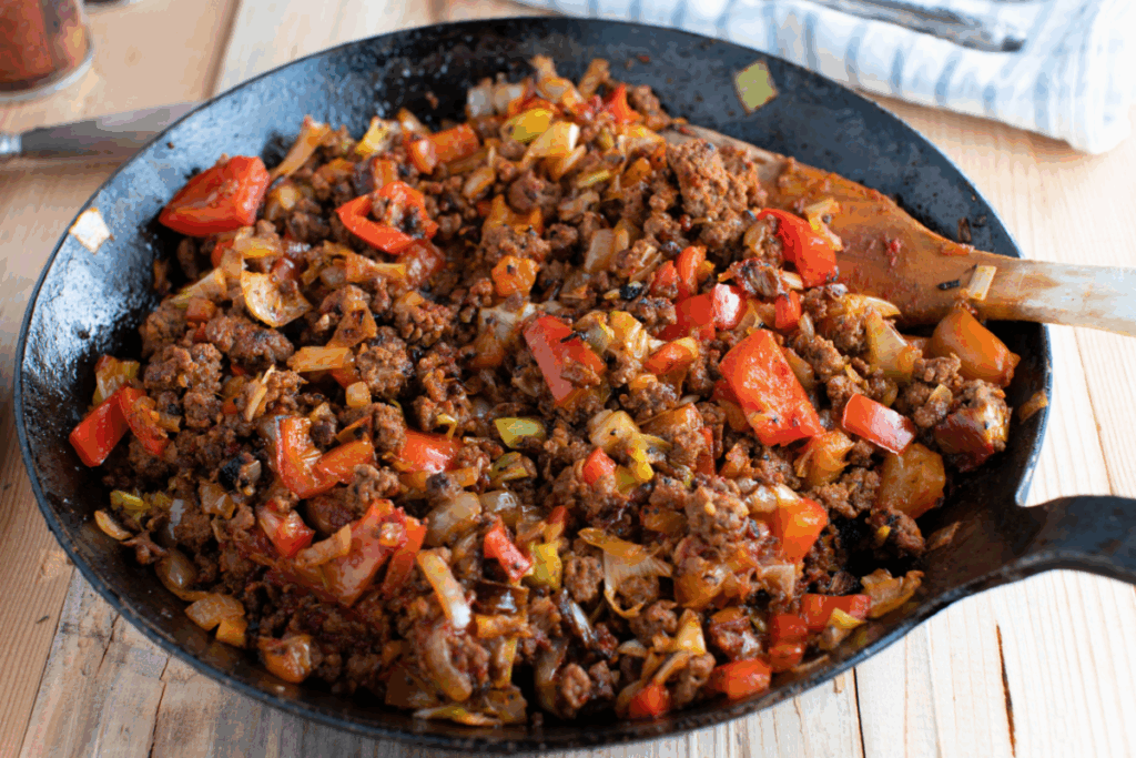 Carne moída refogada com pedaços de pimentão vermelho, cebola e outros vegetais, preparada em uma frigideira de ferro fundido preta que está sobre uma superfície de madeira clara, com uma colher de pau inserida na mistura.