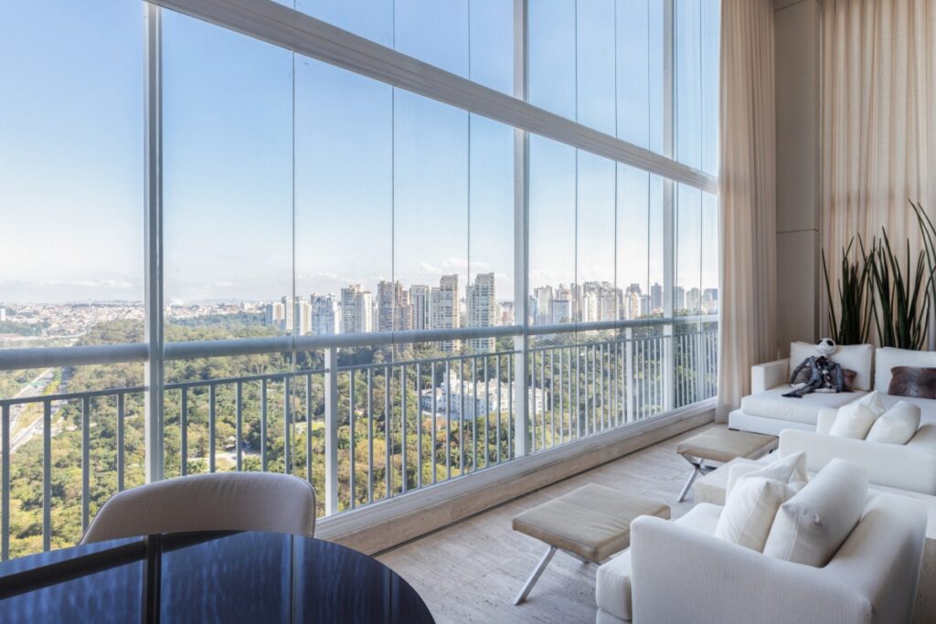 Uma ampla varanda de apartamento com fechamento em vidro, decorada com sofás brancos e oferecendo uma vista panorâmica de uma grande área verde e prédios ao fundo.