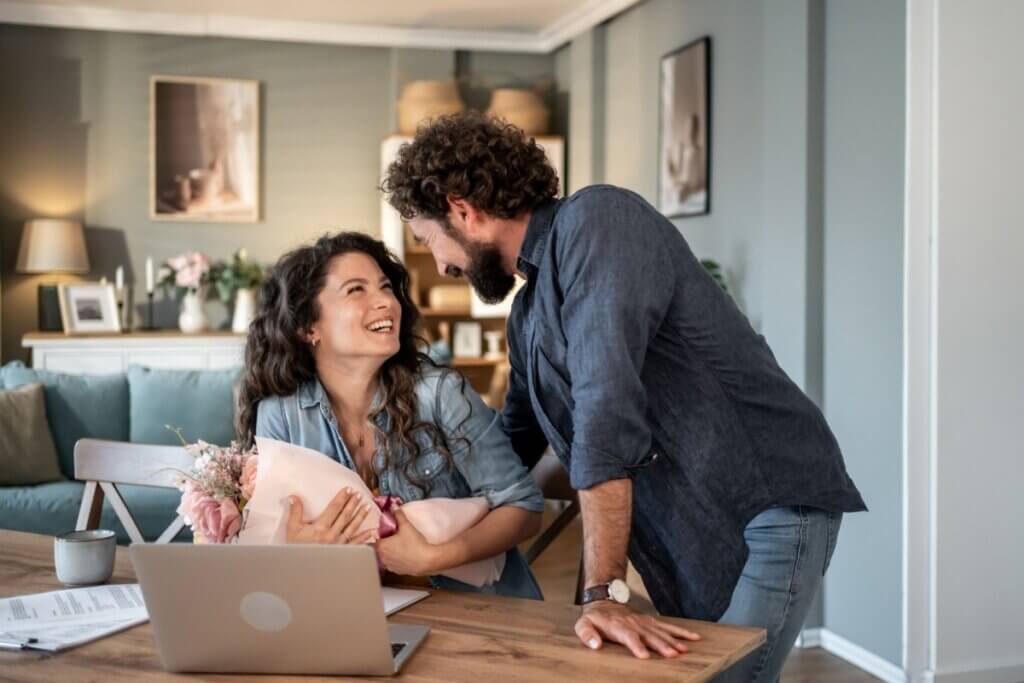 Homem com cabelo cacheado e barba entrega um buquê de flores para uma mulher sentada em frente a um notebook. Ambos sorriem e se olham com carinho em um ambiente residencial aconchegante.