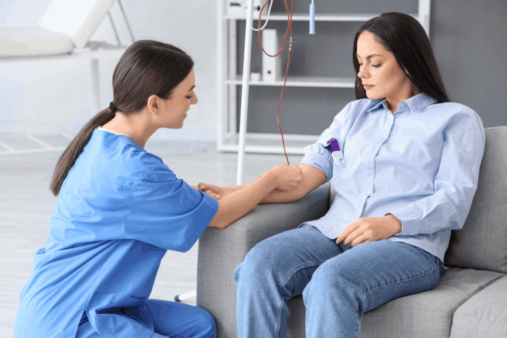 Uma profissional de saúde com uniforme cirúrgico azul, agachada ao lado de uma paciente sentada em um sofá cinza, ajustando ou preparando o braço dela para um procedimento de doação de sangue, conectado a um equipo com tubo vermelho.