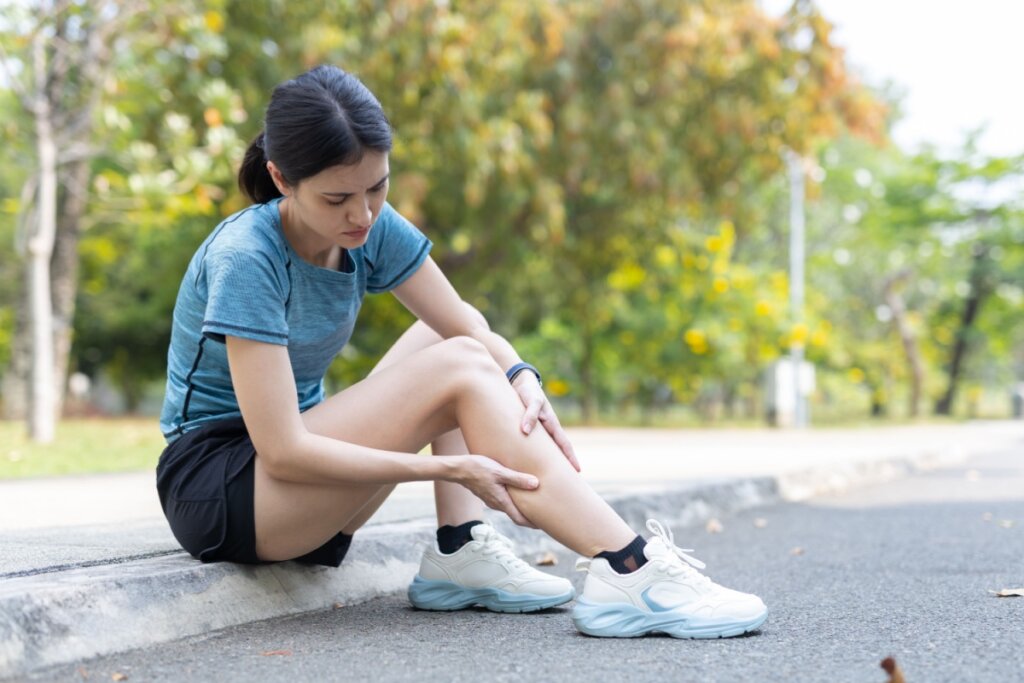 Uma jovem mulher asiática está sentada no meio-fio de uma rua ou pista de corrida, expressando dor. Ela veste uma camiseta azul e shorts pretos. Com as duas mãos, ela segura a perna esquerda na região abaixo do joelho (canela/panturrilha), com o corpo levemente inclinado para a frente e uma expressão facial de desconforto. O fundo mostra um parque com árvores e vegetação levemente desfocadas.