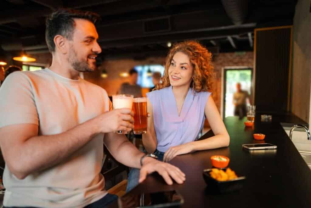 Homem e mulher sorriem e brindam com copos de cerveja no balcão de um bar. O homem usa camiseta clara e a mulher, com cabelos cacheados, veste uma blusa lilás sem mangas. Pequenas porções de petiscos estão espalhadas sobre o balcão escuro.
