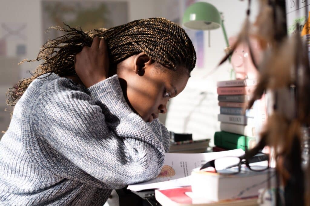 Uma jovem estudante negra, com tranças estilo box braids loiras e castanhas, concentrada na leitura de um livro didático aberto. Ela está sentada à sua mesa, inclinada para a frente, com a mão direita tocando o cabelo na nuca, pensativa. A mesa está repleta de materiais de estudo, incluindo uma pilha de livros coloridos, óculos de leitura sobre uma capa vermelha, uma luminária de mesa verde clara e um cofrinho rosa em forma de porco.