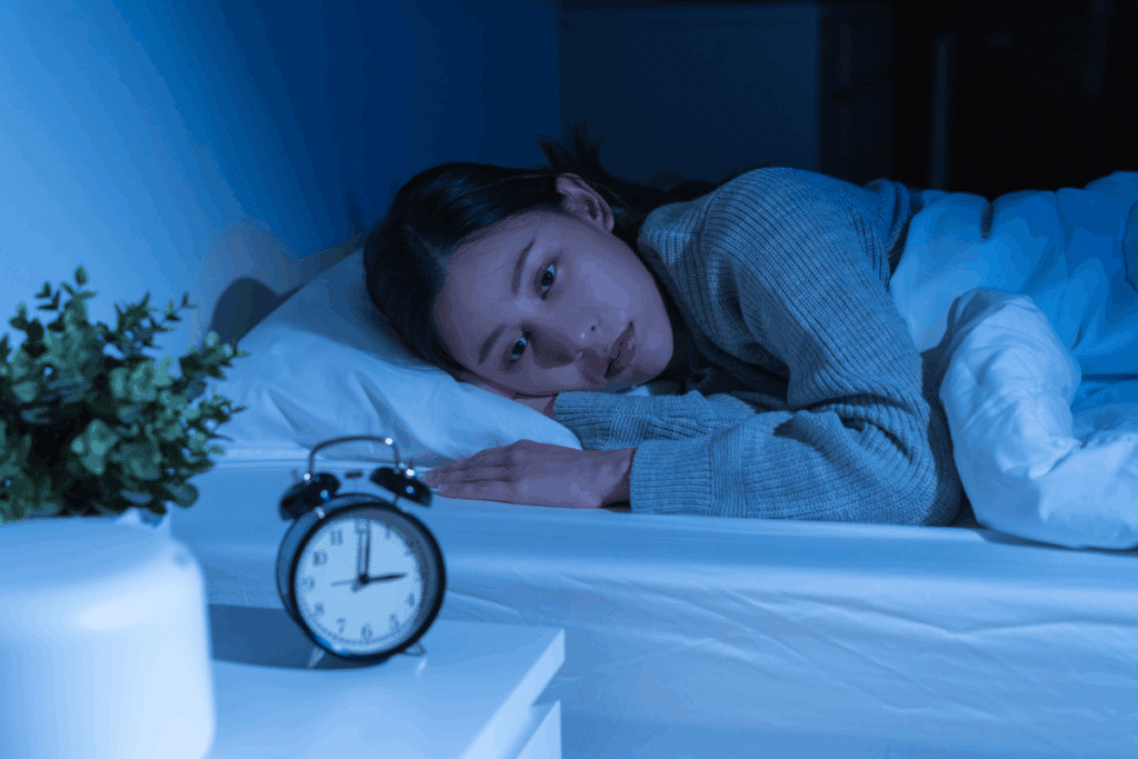 Mulher deitada na cama no escuro, iluminada por uma luz azulada, com os olhos bem abertos e expressão de insônia, enquanto um despertador em primeiro plano marca três horas da manhã.