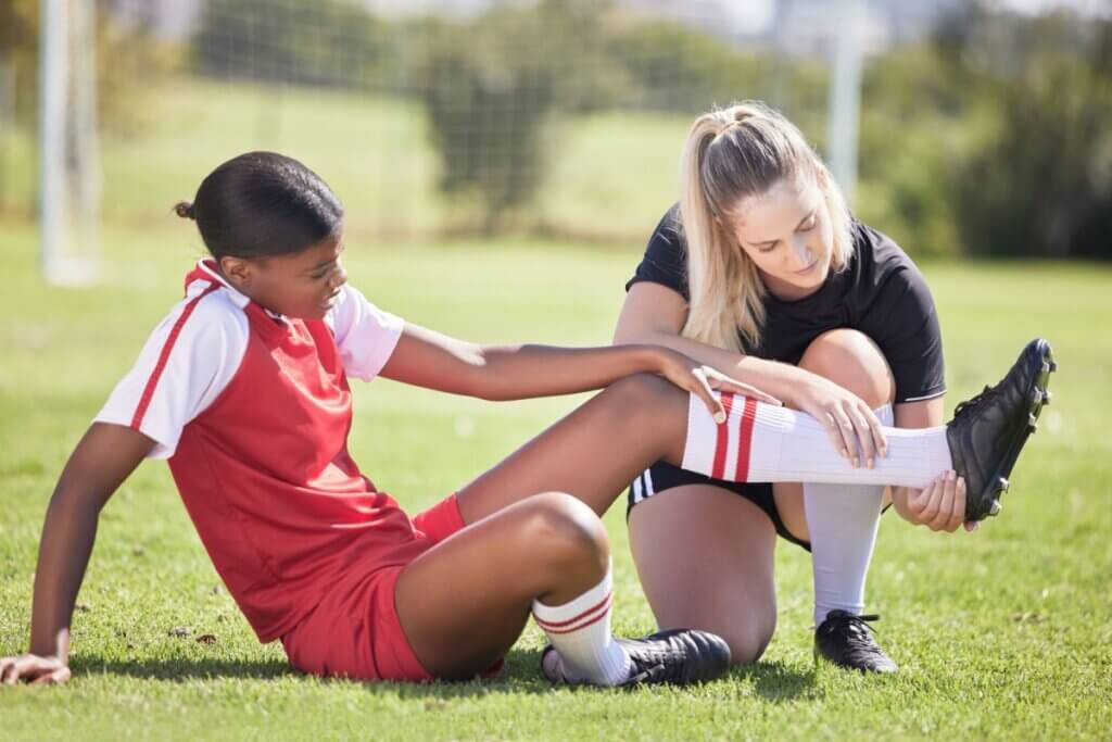 Fotografia em plano médio focada em duas jogadoras de futebol em um gramado. À esquerda, uma jovem atleta negra veste um uniforme vermelho e branco e está sentada no chão com uma expressão de dor ou desconforto, apoiando a mão sobre o próprio joelho. À direita, uma mulher loira, vestindo uniforme totalmente preto, está ajoelhada e segura a perna estendida da jogadora ferida pelos tornozelos, ajudando-a a alongar ou avaliando a lesão. O fundo mostra o restante do campo esportivo de forma desfocada.
