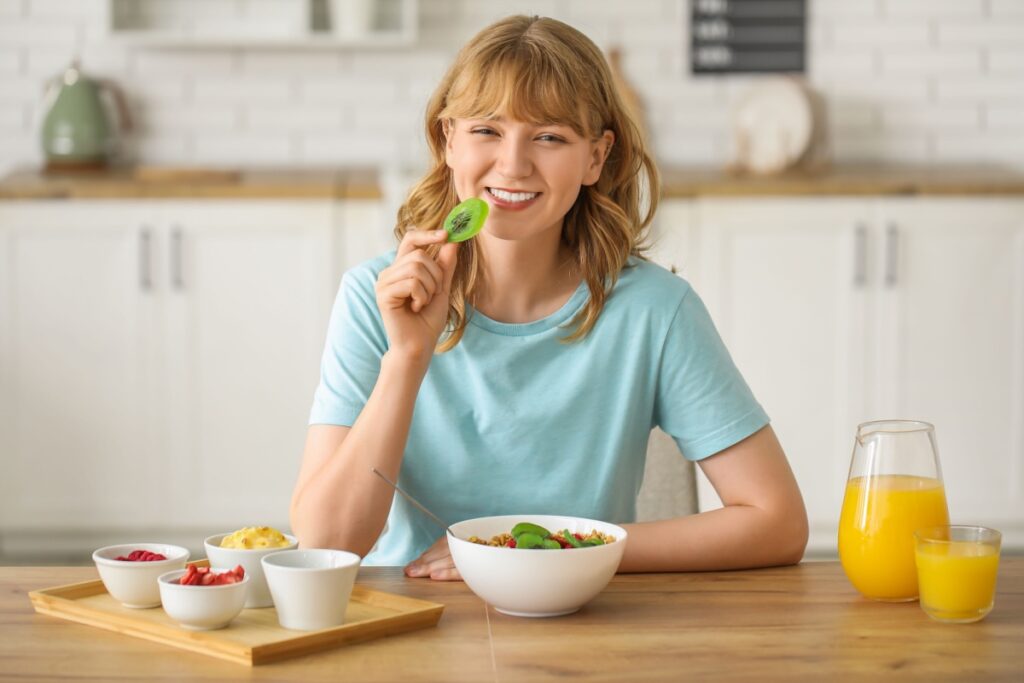 Uma mulher jovem e sorridente, com cabelos loiros e vestindo uma camiseta azul-clara, está sentada à mesa da cozinha tomando café da manhã. Ela segura uma fatia de kiwi perto do rosto. À sua frente, há uma tigela com cereal e frutas, uma jarra e um copo de suco de laranja, além de uma bandeja com pequenos potes contendo acompanhamentos.