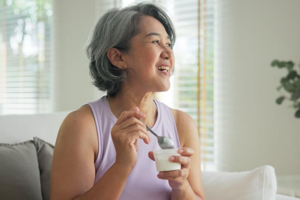 Uma mulher de meia-idade com traços asiáticos e cabelos curtos e grisalhos aparece sorrindo e olhando para o lado direito da imagem. Ela está sentada em um sofá branco, veste uma blusa regata lilás e segura um pote pequeno de iogurte branco em uma das mãos, enquanto usa uma colher com a outra mão. O ambiente ao fundo é uma sala iluminada pela luz natural que entra através de uma janela com persianas.