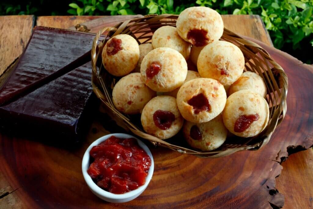Uma cesta de vime cheia de pães de queijo redondos, recheados com goiabada cremosa que aparece no topo de cada um. Ao lado, há um bloco de goiabada cascão e um pequeno pote com mais goiabada pastosa, dispostos sobre uma tábua de madeira rústica.