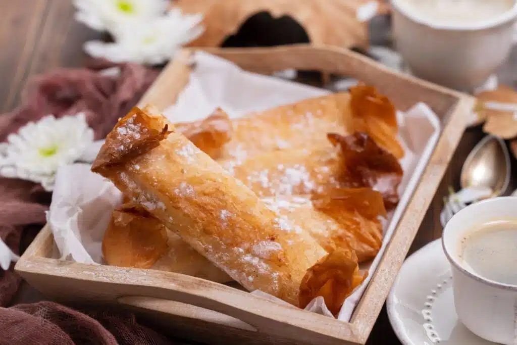 Uma bandeja de madeira forrada com papel branco contém vários Pastéis de Tentúgal, que são doces cilíndricos de massa folhada dourada e crocante salpicados com açúcar de confeiteiro. Ao fundo, há detalhes decorativos com flores brancas e, em primeiro plano à direita, uma xícara de café.