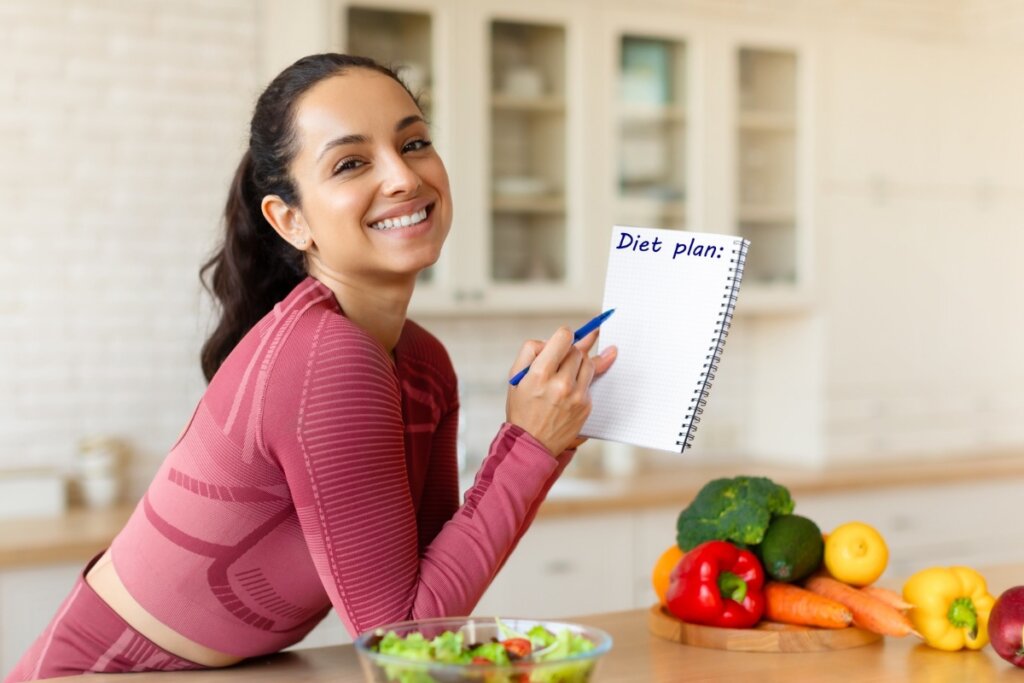 Mulher sorrindo escorada em uma bancada na cozinha segurando um caderno