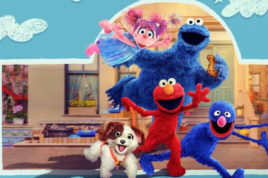 Imagem promocional colorida com fundo azul claro e nuvens desenhadas. No centro, os fantoches Elmo (vermelho), Come-Come (azul, segurando um biscoito), Abby Cadabby (rosa, voando com varinha de condão), Grover (azul escuro) e um pequeno cachorro branco e marrom posam sorridentes e acenando em frente ao cenário da Vila Sésamo.