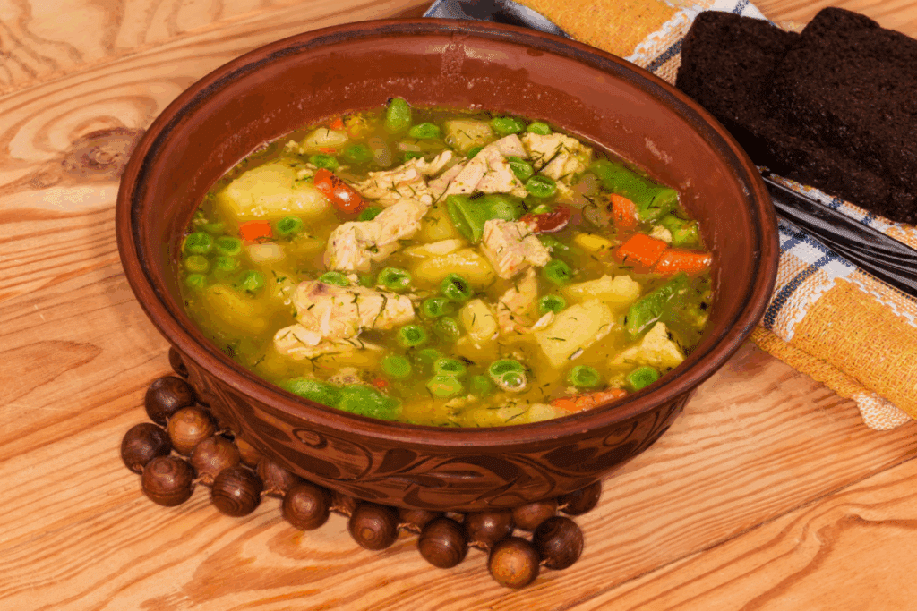 Imagem mostrando uma tigela de cerâmica marrom cheia de sopa de frango em cubos com legumes visíveis, como ervilhas, cenouras e batatas, salpicada com ervas. A tigela está sobre um descanso de contas de madeira em uma mesa rústica, ao lado de fatias de pão preto.