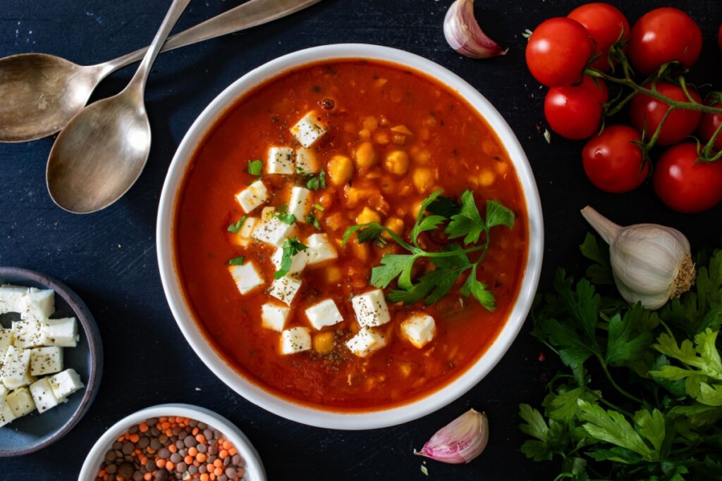 Sopa de lentilha e grão-de-bico com cubos de queijo feta, servida em tigela branca e decorada com folhas de salsa