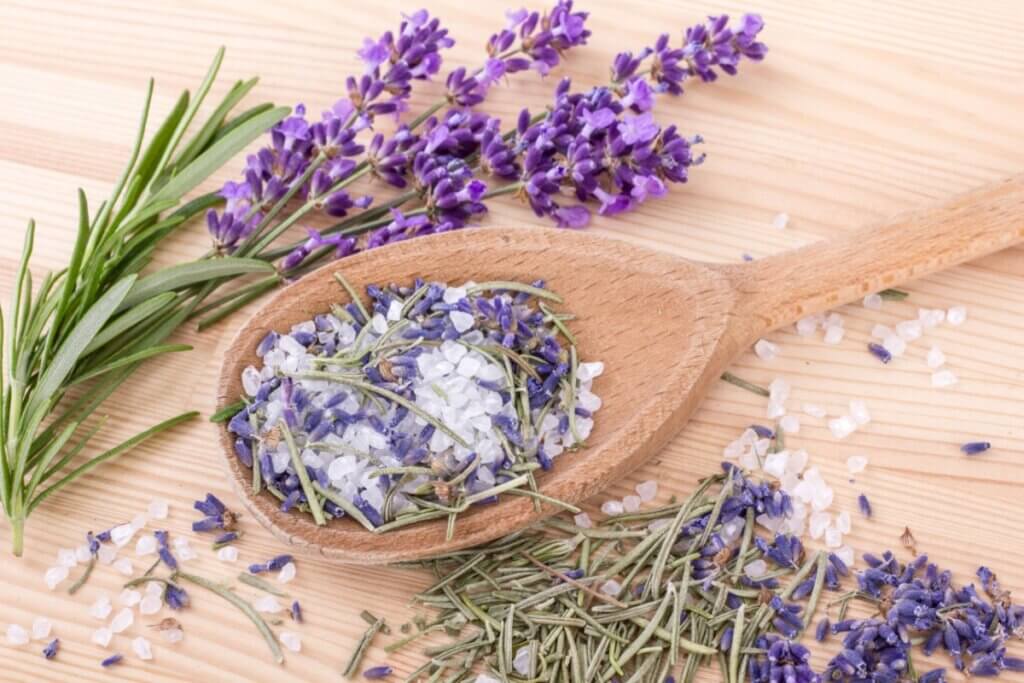 A imagem mostra uma colher de madeira com uma mistura de sal grosso e flores secas, como lavanda e ramos de alecrim. Ao redor, há mais ervas e flores espalhadas sobre uma superfície de madeira clara.