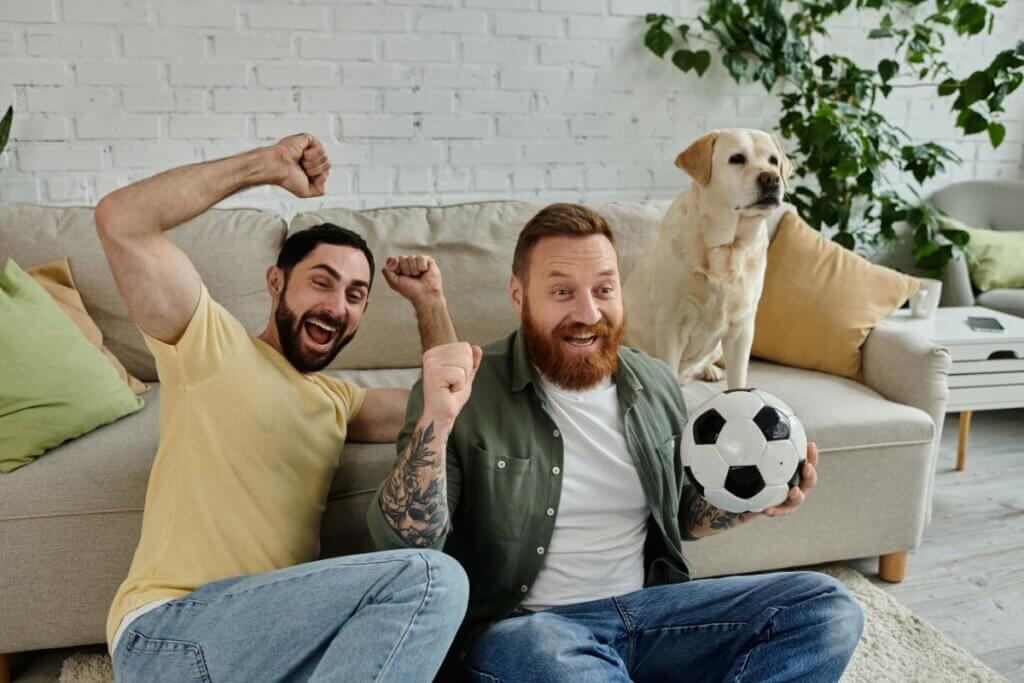 Dois homens, sentados no chão e assistindo futebol, cachorro sentado no sofá ao lado