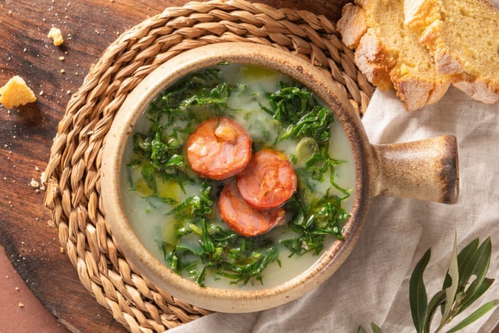 Caldo verde servido em panela com couve fatiada e rodelas de linguiça, acompanhado por fatias de pão sobre uma mesa
