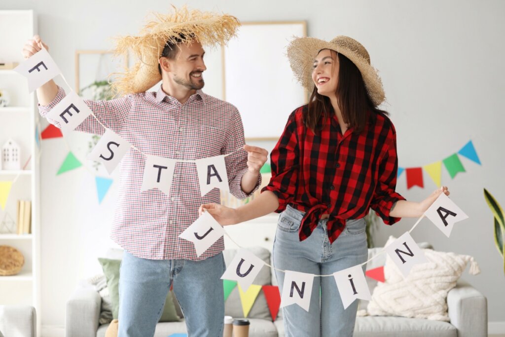 Casal vestido com camisas xadrez e segurando uma corrente de bandeirinha escrito festa junina e mais bandeirinhas atrás na parede da sala
