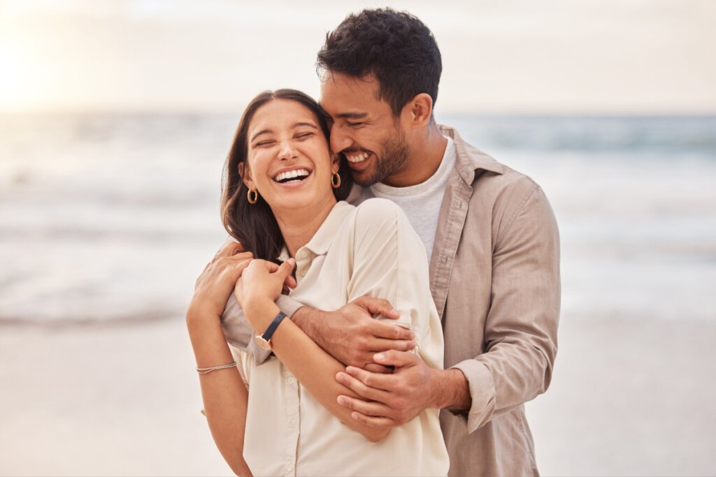 Mulher usando camisa bege, com o cabelo solto, abraçada com homem com cabelo curto e barba usando camisa bege e camiseta branca. Ambos estão sorrindo