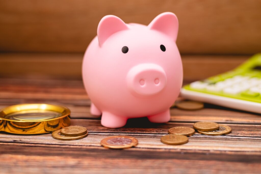cofrinho de porquinho rosa com moedas em volta, calculadora verde e branca ao fundo e lupa amarela ao lado