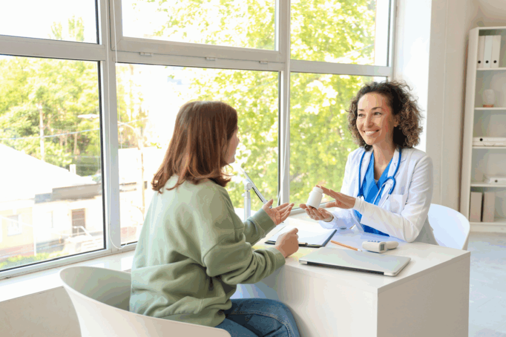 médica e paciente sentadas uma de frente para a outra. médica segura frasco de remédio e sorri. Paciente tem o cabelo claro, curto e está usando camisa verde e calça jeans e médica tem o cabelo curto cacheado e está usando jaleco branco e camisa azul com estetoscópio no pescoço